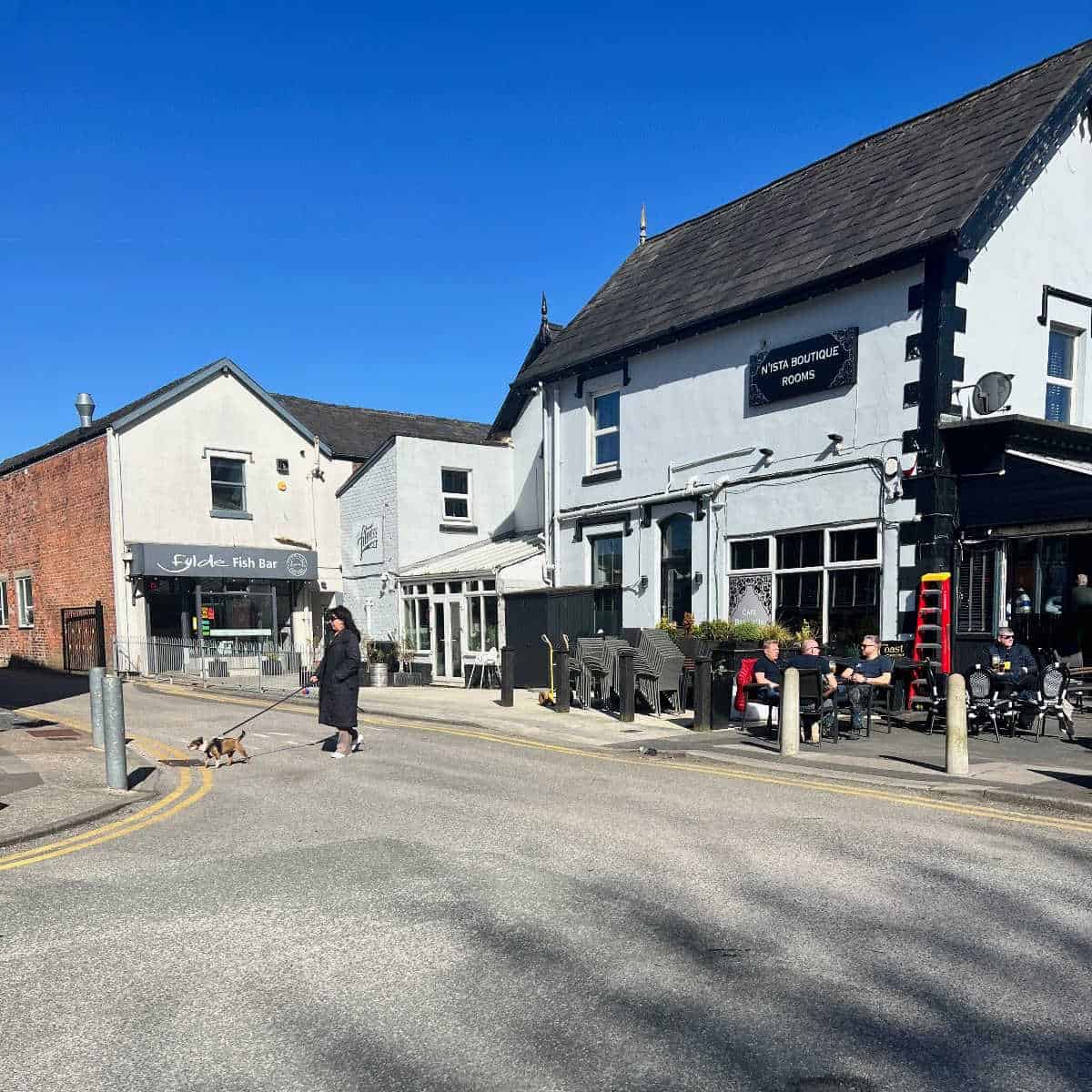 Street view of Birkdale, showing men enjoying a drink outside of a cafe in Birkdale and a lady wearing a black coat and sunglasses walking her dog